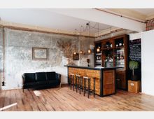 Eine rustikale Bar in einem Raum mit sichtbaren Putzwänden, Holzfußboden und Stuckdecke. Die Bar besteht aus einer hölzernen Schrankfront mit kleinen Schubladen und ist von Barhockern umgeben; daneben befindet sich ein Regal mit Gläsern und Flaschen. Links davon steht ein schwarzes Ledersofa an der Wand.