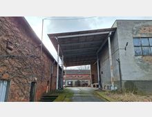 Überdachter Durchfahrtsbereich zwischen einem roten Backsteingebäude und einem grauen Industriegebäude mit großen Fenstern und bröckelndem Putz. Betondecke auf Stahlstützen über einer teils vermoosten Betonfläche; im Hintergrund weitere Betriebsgebäude und eine Mauer mit Graffiti.