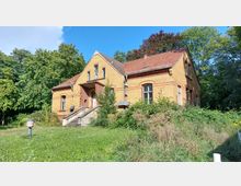 Backsteingebäude mit rotem Ziegeldach, gelegen in einer grünen, bewachsenen Umgebung. Das einstöckige Gebäude hat eine Treppe mit Eingangsbereich sowie mehrere große Fenster und steht inmitten von Gras und Sträuchern, umgeben von Bäumen.