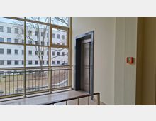 Innenflur eines modernen Gebäudes mit Aufzugtür in einem dunklen Rahmen und großflächigen Fensterfronten. Vor den Fenstern verläuft ein Metallgeländer; draußen sind ein Innenhof mit Bäumen und ein gegenüberliegendes mehrstöckiges Gebäude zu sehen.