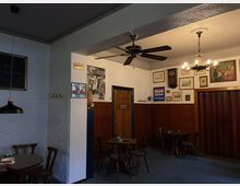 Gemütlicher Gastraum mit Holzmöbeln, vertäfelten Wänden und dekorativen Elementen wie Bildern, einer antiken Wanduhr und einem Wandventilator. Die Beleuchtung besteht aus einem Kronleuchter und Hängelampen, und eine Tür mit der Aufschrift „Toilette“ ist sichtbar.