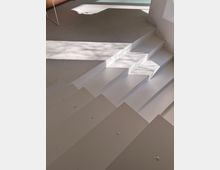 Interior view of a modern space with a wide white staircase leading up to a speckled terrazzo-like floor. Sunlight casts sharp rectangular shadows across the steps and landing near a white wall.