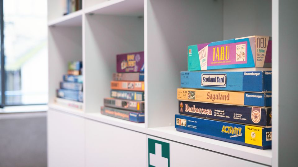 Weiße Regale in einem hellen Innenraum, mit mehreren gestapelten Brettspielen in den Fächern. Unten ist ein Schrank mit einem grünen Erste-Hilfe-Symbol zu sehen, daneben ein großes Fenster im Hintergrund.