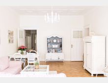 Helles Wohnzimmer mit Holzparkettboden und weißer Einrichtung im Vintage-Stil, darunter ein Schrank, ein Regal mit Dekorationen sowie ein Esstisch und Stühle. Eine weiße Hängelampe und zart rosafarbene gestreifte Polstermöbel ergänzen die Einrichtung.