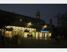Bahnhof Berlin-Spandau bei Nacht mit glasüberdachtem Eingangsbereich, beleuchteten Schaufenstern und Schildern von Geschäften wie „Sparda-Bank“ und der Deutschen Bahn. Im Hintergrund ist ein Uhrturm und im Vordergrund ein Vorplatz mit parkenden Fahrrädern und wenigen Personen.