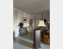 Ein modernes Wohnzimmer mit minimalistischer Einrichtung, grauen Wänden und einem hellen Boden. Links steht ein Regal mit Büchern und Schallplatten, darüber hängt ein gerahmtes Kunstwerk. Im Vordergrund sind eine Stehlampe, ein Lautsprecher und ein senfgelbes Sofa zu sehen.
