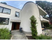 Zweistöckiges Wohnhaus mit weißer Fassade und abgerundeten Wandelementen, großen rechteckigen Fenstern und einem kleinen Vordach über dem Eingang. Im Vordergrund ein gepflasterter Weg, ein Fahrrad, grüne Pflanzen und im Hintergrund eine Terrasse mit Gartenmöbeln.