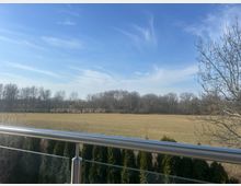 Blick von einem Balkon mit Glasgeländer über eine große, flache Wiese bis zu einer Baumreihe am Horizont. Im Vordergrund stehen dichte Hecken, darüber ein weiter blauer Himmel mit dünnen Wolken.