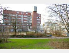 Backstein-Gebäude mit mehreren Stockwerken und zahlreichen Fenstern, umgeben von kahlen Bäumen in einem parkähnlichen Gelände mit Rasenflächen und Wegen. Im Hintergrund sind weitere Gebäude und Graffiti an Mauern sichtbar.