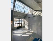 Innenraum eines loftartigen Gebäudes mit hohen Decken, einer großen Glaswand und Oberlichtern, die Tageslicht hereinlassen. Der Raum hat graue Wände, eine schlichte Betonoptik und moderne Hängelampen.