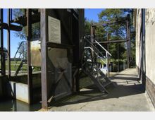 Industrielles Schleusenbauwerk mit Metalltreppen, Drehgriffen und einer informativen Hinweistafel. Im Hintergrund sind ein kleiner Fluss, Wiesen und Bäume bei sonnigem Wetter zu sehen.