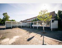 Eine Außenansicht mit einem gepflasterten Platz und mehreren weißen, überdachten Containern, die als Sitzbereiche und Verkaufsstände genutzt werden. Im Hintergrund ist ein langgezogenes Backsteingebäude mit großen Fenstern sowie einige Bäume sichtbar.