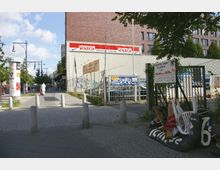 Straßenansicht in einer urbanen Umgebung mit Pflasterweg und Pollern, flankiert von Bäumen und einem Werbesäulenplakat. Rechts ist ein eingezäuntes Gelände mit geparkten Autos und einem Gebäudekomplex aus rotem Backstein im Hintergrund sichtbar.