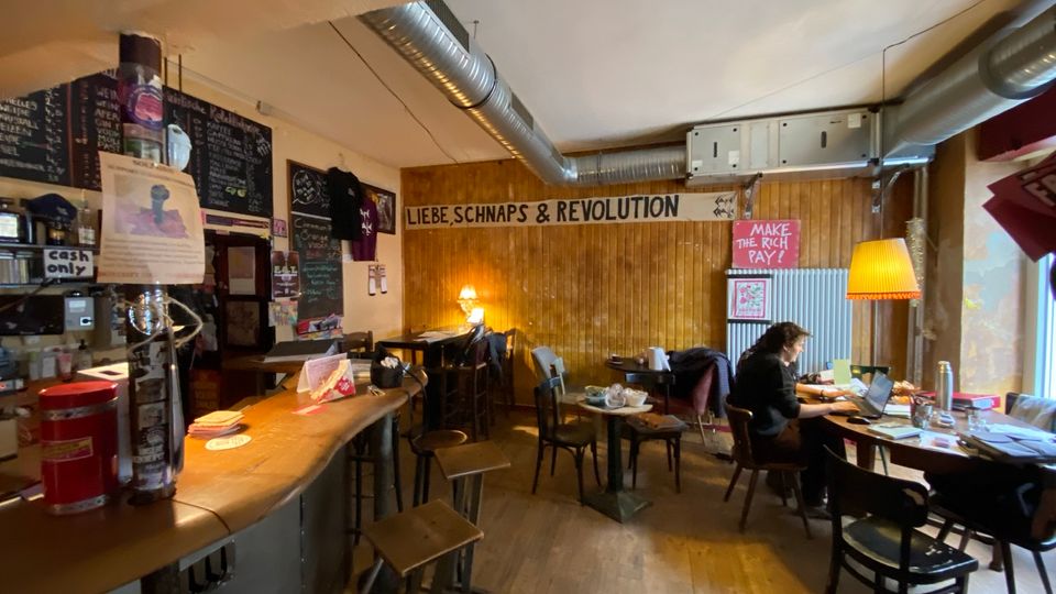 Innenraum einer gemütlichen Bar oder eines Cafés mit langer Theke links, Holzwand und mehreren kleinen Tischen und Stühlen auf Holzboden. Sichtbare Lüftungsrohre an der Decke, Wandtafeln mit Speisekarte und politische Poster, dazu warme Lampenbeleuchtung.