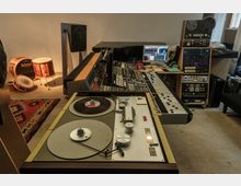 Tonstudio mit analoger und digitaler Ausrüstung: Im Vordergrund ein Tonbandgerät, dahinter ein großes Mischpult mit Reglern und Knöpfen. Im Hintergrund befinden sich Lautsprecher, ein Stapel Audiogeräte, Computerbildschirm und mehrere Schlaginstrumente in einem schallgedämpften Raum.