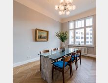 Esszimmer mit einem Glastisch auf einem Steinsockel, umgeben von sechs Holzstühlen mit blauen Sitzpolstern, auf einem Fischgrät-Parkettboden. Ein großes Fenster mit Blick auf moderne Gebäude und eine Blume in einer Vase sowie ein gerahmtes Gemälde schmücken den Raum.