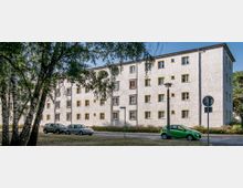 Wohnblock in schlichter Bauweise mit weißer Fassade, gelben Fensterrahmen und vier Etagen. Im Vordergrund stehen geparkte Autos sowie vereinzelte Bäume, die das Bild leicht verschatten.