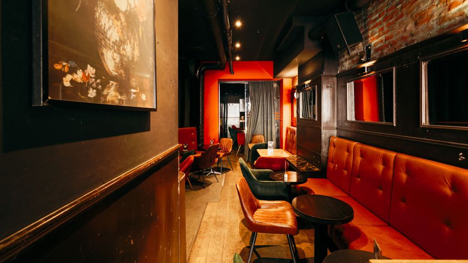 Schmaler Bar- oder Loungebereich mit roten gepolsterten Sitzbänken, kleinen runden Tischen und gemischten Stühlen entlang eines Holzfußbodens. Dunkle Decke mit sichtbaren Rohrleitungen und Spotbeleuchtung, rechts eine freiliegende Ziegelwand mit Spiegeln und am Ende ein rot gerahmter Durchgang mit Vorhang.
