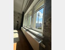 Innenansicht eines Raumes mit einem großen Fenster, das den Berliner Fernsehturm und einen Teil der Stadtlandschaft zeigt. Der Raum hat einen Holzboden, eine Heizung unter dem Fenster und eine gemusterte Wandtapete.