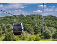 Seilbahn mit modernen Gondeln, die über eine grüne Parklandschaft mit Bäumen und Wiesen führt; im Hintergrund bewaldete Hügel und ein weißes Gebäude auf einer Anhöhe.