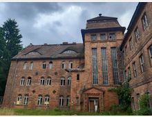 Ehemaliges Backsteingebäude mit mehreren Etagen und hohen, schmalen Fenstern, teilweise beschädigt und von Vegetation umgeben. Der Himmel ist bewölkt, und das Gebäude wirkt verlassen und ungenutzt.