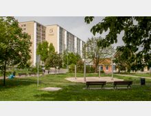 Park mit Rasenfläche, Bänken und blühenden Bäumen im Vordergrund; im Hintergrund ein großes Wohngebäude mit mehreren Etagen sowie ein flacheres rotes Gebäude. Spazierwege und vereinzelte Laternen sind sichtbar.