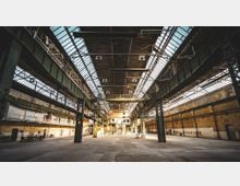 Weitläufige Industriehalle mit hohen Decken aus Stahlträgern und Glasfenstern, die viel Tageslicht einlassen. Rechts und links befinden sich backsteinverkleidete Wände, und zentrale Stahlträger tragen die Struktur.