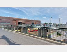 Bahnhof Warschauer Straße in Berlin, mit einem modernen, flachen Gebäude aus braunem Metall im Hintergrund, einer erhöhten Fußgängerbrücke im Vordergrund und einem darunter verlaufenden Gleisbereich mit vorbeifahrenden S-Bahn-Zügen.