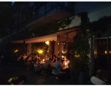 Außenbereich eines Restaurants oder Cafés bei Nacht, mit Tischen voller Gäste, die bei Kerzenlicht sitzen. Die Fassade ist mit Pflanzen und Lichtern dekoriert, und ein Vordach überspannt den Eingangsbereich.