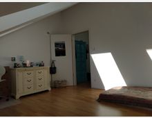 Ein schlichtes Zimmer mit weiß gestrichenen Wänden, einer schrägen Decke und Holzboden. Es enthält eine cremefarbene Kommode, ein blaues Holzschrank, ein flaches Matratzenlager am Boden und eine Tür, die in einen angrenzenden Raum führt; Licht fällt durch ein Fenster in das Zimmer.