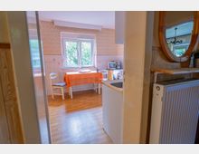Blick in eine kleine Wohnküche mit Holzfußboden, gemusterter Tapete und einem Fenster über einem Tisch mit orangefarbener Tischdecke und zwei Stühlen. Rechts sind Küchenzeile mit Spüle sowie ein Regal mit Spiegel und einem Heizkörper zu sehen.