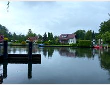 Ein ruhiger Fluss mit spiegelnder Wasseroberfläche, umgeben von kleinen Einfamilienhäusern mit roten Dächern und gepflegten Gärten. Am Flussufer sind ein Bootssteg, ein blaues Boot und ein rotes Ausflugsschiff zu sehen, mit Bäumen und Sträuchern im Hintergrund.