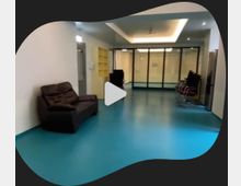 Ein heller Innenraum mit glattem, türkisfarbenem Boden, weißen Wänden und einer abgehängten Decke mit indirekter Beleuchtung. Links steht ein braunes Sofa, rechts sind gestapelte Stühle, und im Hintergrund befinden sich eine große Glaswand und Türen.