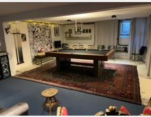 Ein Hobbyraum mit einem Billardtisch auf einem großen, gemusterten Teppich. An der linken Wand befinden sich Wanddeko, ein Gestell mit Billardstöcken und ein Lautsprechersystem, während im Hintergrund Stühle und eine gemusterte Wandzeichnung zu sehen sind.