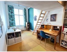 Ein kleines Zimmer mit einem Hochbett, erreichbar über eine Holzleiter. Darunter befindet sich ein Schreibtisch aus Holz mit einer Schreibtischlampe und Regalfächern. Links im Raum steht ein Sofa mit einem Bücherregal davor und einem großen Fenster mit bunten Vorhängen, das viel Tageslicht hereinlässt.