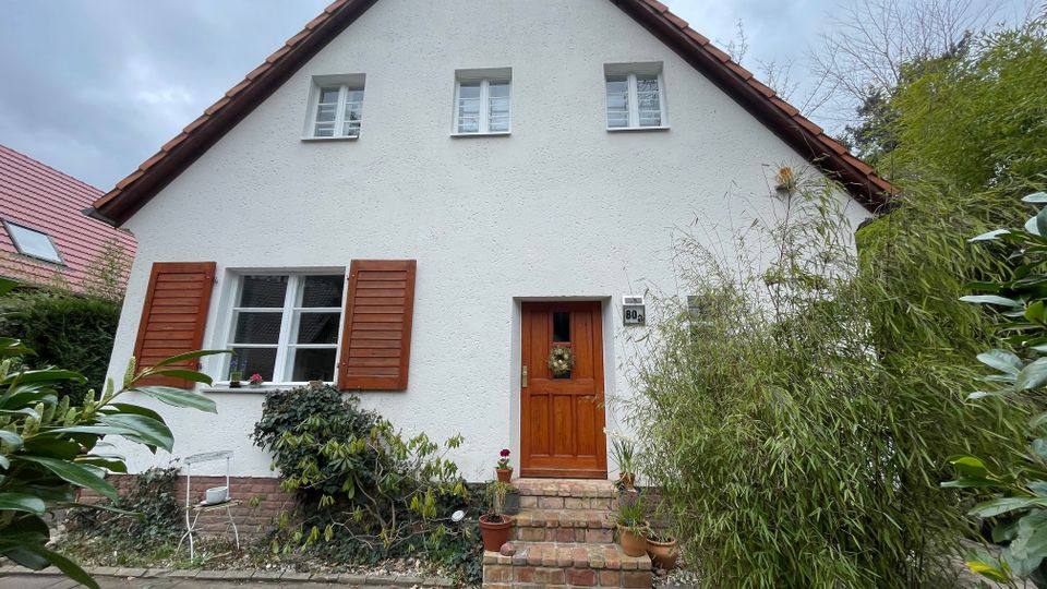 Frontansicht eines kleinen Einfamilienhauses mit weiß verputzter Fassade und steilem Satteldach mit roten Ziegeln. Zu sehen sind Holzfensterläden, eine hölzerne Eingangstür mit kurzen Backsteinstufen sowie ein Vorgarten mit Sträuchern, Bambus und Pflanzkübeln entlang eines gepflasterten Weges.