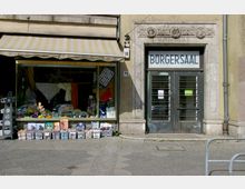 Fassade eines Gebäudes mit zwei Bereichen: links ein Laden mit Schaufenster, in dem verschiedene Waren wie Garne und Dekorationsartikel ausgestellt sind, rechts doppelflügelige Glastüren mit der Aufschrift "Bürgersaal" und dekorativen Steinreliefs über dem Eingang.