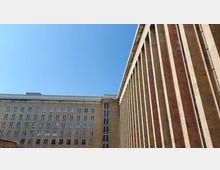 Großes, monumentales Gebäude mit sandfarbener Fassade und zahlreichen rechteckigen Fenstern; die rechte Seite des Bildes zeigt eine Fassade mit vertikalen Säulen. Der Himmel ist klar und blau.