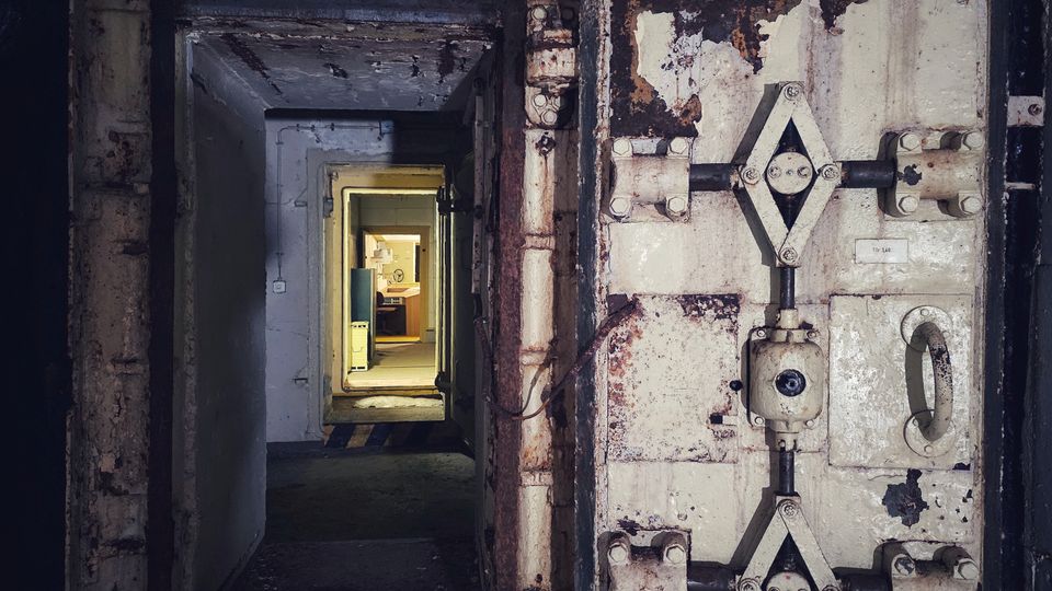 Blick in einen schmalen, betonierten Gang mit einer geöffneten, schweren Stahltür mit rostigen Beschlägen und Verriegelungsmechanismus, typisch für einen Bunker oder Schutzraum. Im Hintergrund führt der Korridor durch weitere Türrahmen zu einem helleren Raum.