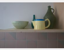 Ein schlichtes Holzregal mit einer pastellfarbenen Keramikschale, einer gelben Teekanne mit blauem Deckel und zwei grünen Vasen. Im Hintergrund sind beige Fliesen mit geometrischen Mustern sichtbar.