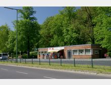 Ziegelgebäude mit flachem Dach, das als kleiner Kiosk genutzt wird, daneben ein S-Bahn-Schild. Im Vordergrund stehen Radfahrer auf einem Gehweg, dahinter befinden sich Grünflächen und Bäume.