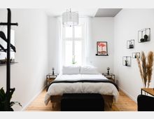 Minimalistisch eingerichtetes Schlafzimmer mit einem Doppelbett in der Mitte, flankiert von zwei schwarzen Nachttischen. Große Fenster mit weißen Vorhängen sorgen für viel Tageslicht, an der Wand hängen Regale mit Pflanzen und ein gerahmtes Flamingo-Poster.