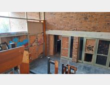 Innenraum eines verlassenen Gebäudes mit hohen Ziegelwänden und großen Fensterflächen; mehrere Türen und Fenster sind mit Sperrholzplatten vernagelt und mit Graffiti beschmiert. Im Vordergrund stehen beschädigte Holzbänke auf einem staubigen Boden.