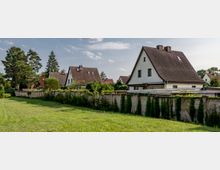 Reihen von Einfamilienhäusern mit spitzen Dächern und weißen Fassaden, dahinter eine bewachsene Mauer und eine Rasenfläche im Vordergrund; umgeben von Bäumen und Büschen.