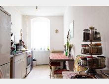 Schmale Küche mit einer Arbeitszeile links, ausgestattet mit Herd, Spüle und Küchenutensilien. Rechts ein Esstisch mit gemusterter Tischdecke, Blumenvasen und Sitzgelegenheiten; im Vordergrund ein drehbares Regal mit Gewürzgläsern.