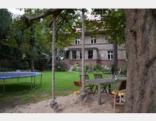 Ein Garten mit einem großen Baum im Vordergrund, der eine Schaukel mit dicken Seilen trägt. Im Hintergrund steht ein zweistöckiges Haus mit Putz- und Backsteinfassade, umgeben von gepflegtem Rasen, Blumenbeeten und Gartenmöbeln auf einem Sandbereich.