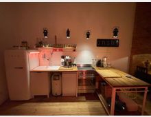Kleine, gemütliche Küche mit weißen Wänden und Holzboden. Ausgestattet mit einem weißen Retro-Kühlschrank, einer Spüle, einem Elektroherd mit Backofen und offenen Holzarbeitsflächen; darüber befinden sich Gewürzregale und hängende Lampen.