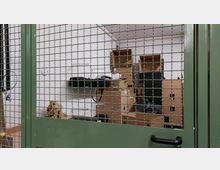 Raum mit weiß gestrichenen Wänden hinter einem grünen Metallgitter; im Inneren befinden sich gestapelte Kartons, Regale und verschiedene Gegenstände wie Taschen.