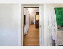 Türöffnung zu einem modernen Wohnbereich mit hellem Holzboden, weißen Wänden und minimalistischem Mobiliar. Rechts im Vordergrund sind ein Handtuchhalter mit grünen Handtüchern und ein aufgehängter Bademantel sichtbar.