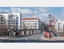 Eine Straße mit einer schmalen Brücke im Vordergrund, flankiert von modernen und historischen Wohngebäuden mit weißen Fassaden. Straßenlaternen und Verkehrsschilder sind sichtbar, darunter ein Schild mit der Aufschrift "Monumentenstraße" und eine Geschwindigkeitsbeschränkung von 30 km/h.